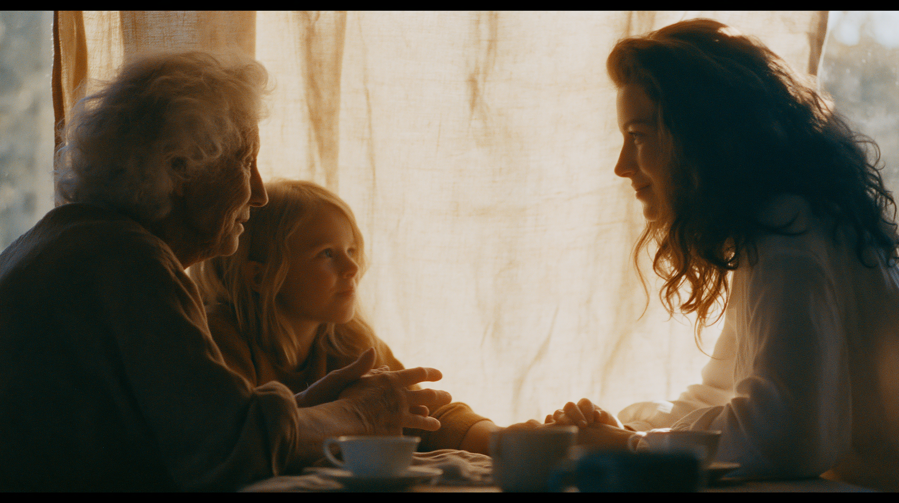 Three generations in unhurried conversation at a sunlit table — grandmother, grandchild and mother holding hands